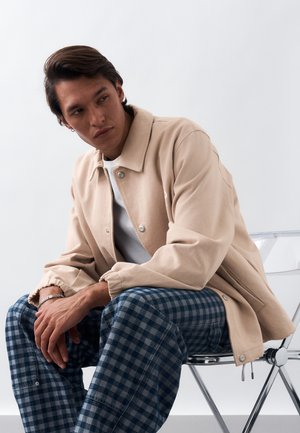 Νέος άνδρας με σκούρα μαλλιά κάθεται σε διαφανή καρέκλα φορώντας μπεζ σακάκι, λευκό πουκάμισο και μπλε παντελόνι με καρό σχεδίαση, πάνω σε απλό φόντο.