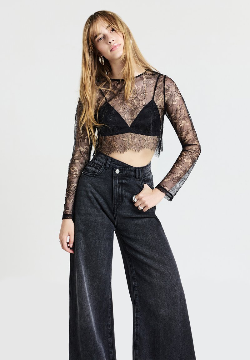 Schwarzes Spitzen-Crop-Top mit langen Ärmeln, durchsichtigen Details und gewelltem Saum, kombiniert mit hoch taillierten, weit geschnittenen schwarzen Jeans.