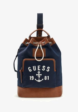 Sac à dos bleu marine et marron avec poche avant zippée, fermeture par cordon de serrage, et texte "GUESS 1981" avec logo ancre sur la poche avant.