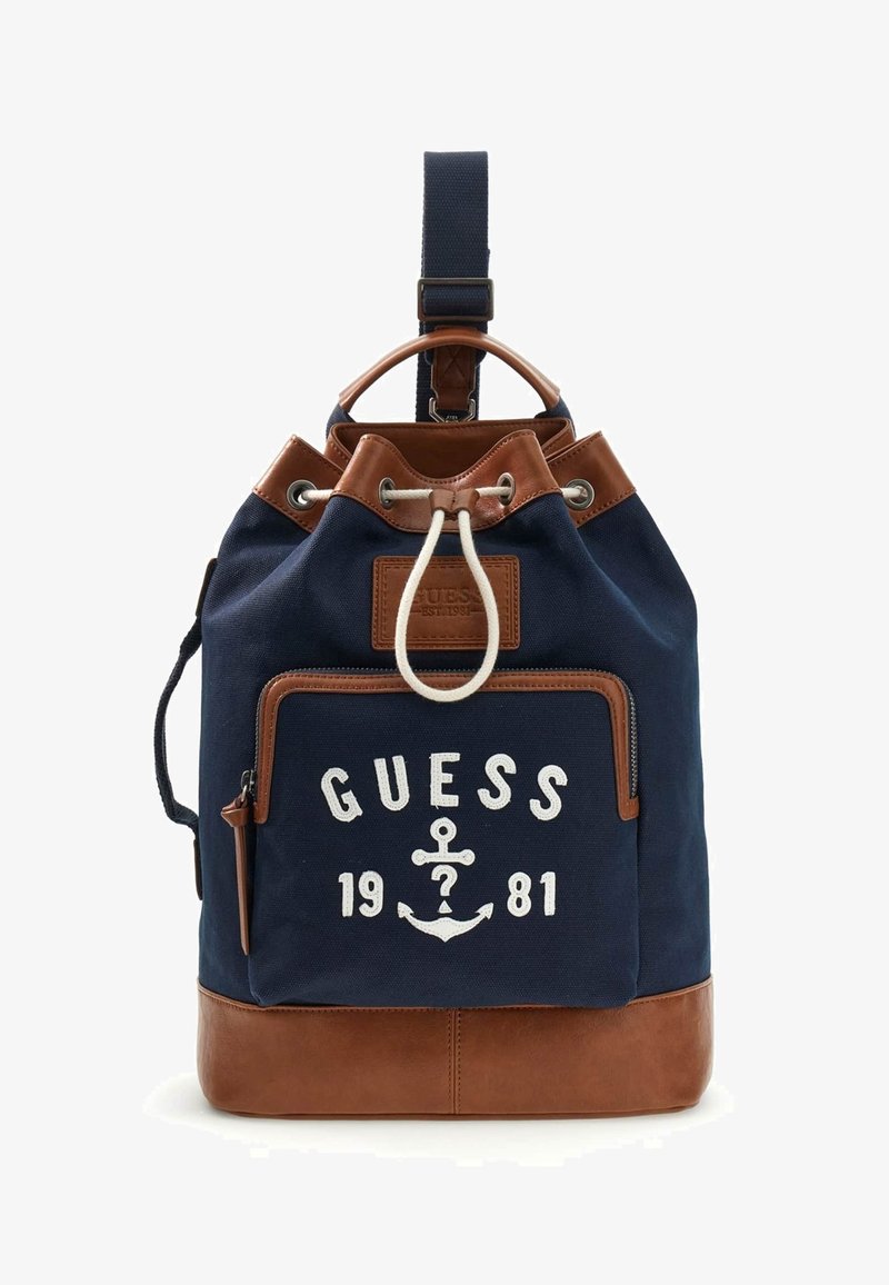 Sac à dos bleu marine et marron avec poche avant zippée, fermeture par cordon de serrage, et texte "GUESS 1981" avec logo ancre sur la poche avant.