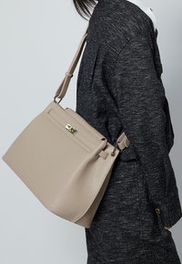 Beige læderhåndtaske med tekstureret overflade, der har en top-håndtag, justerbar rem og en guldtonet lås. Detalje med sidelomme med lynlås.