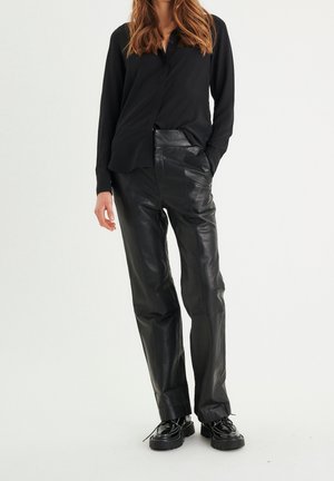 Nainen, joka pukeutunut mustaan napitettuun paitaan, mustiin nahkahousuihin ja mustiin paksupohjaisiin nauhallisiin kenkiin, seisomassa käsi taskussa.