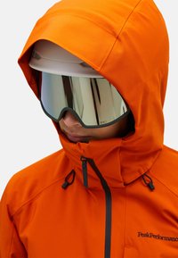 Orange vandtæt jakke med høj krave, justerbar hætte og sort lynlåsdetalje. Reflekterende goggles forbedrer synligheden.