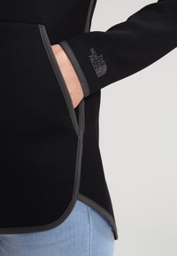 Giacca in pile nera con dettagli grigi, vestibilità aderente e logo visibile sulla manica. Include una tasca laterale per il recupero.