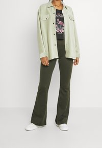 Giacca camicia in corduroy verde chiaro con due tasche sul petto, indossata sopra una t-shirt nera con un grafico che rappresenta un fenicottero. Pantaloni a campana verde oliva e sneakers bianche.