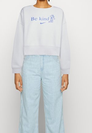 Vrouw draagt een lavendelkleurige trui met de tekst "Wees vriendelijk" en een bloemenafbeelding, gecombineerd met lichtblauwe wijde jeans.