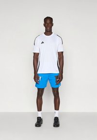 Wit sportief shirt met zwarte strepen, gecombineerd met blauwe shorts met een grafische opdruk. Zwarte sneakers maken de outfit compleet.