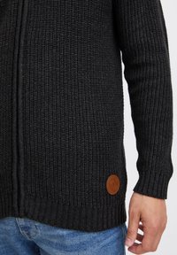 Mustan neulecardigan, jossa on röyhelötekstuuri ja vasemmalla puolella nahkainen logopatchi. Vetoketjullinen malli pitkillä hihoilla.