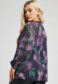 Genomskinlig blus i lila och grön blommönster, med puffärmar och rund halsringning. Strukturerad tyg med subtil lyster och elastiska ärmslut.