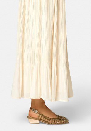 Pies con zapatos slingback beiges con tachuelas y tacón bajo, combinados con una falda larga plisada de color crema.