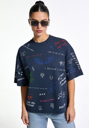 Vrouw met donker haar in een knot, die donkere zonnebril en een marineblauw t-shirt draagt dat bedekt is met kleurrijke handgeschreven tekst en krabbels.