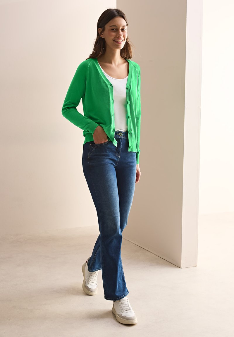 Grön kofta med knappar, vit T-shirt, mörkblå utsvängda jeans och vita sneakers. Slät yta och avslappnad design.