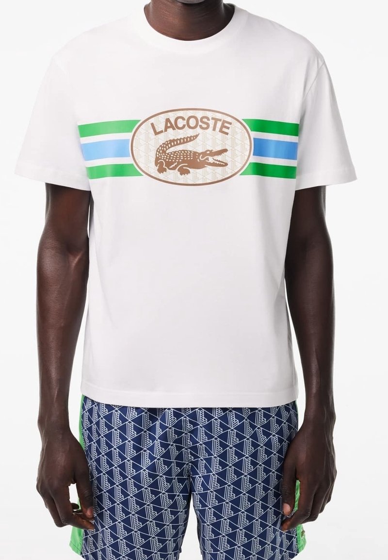 Camiseta de algodón blanca con acentos en rayas verdes y azules y un logo ovalado de Lacoste en marrón con un gráfico de cocodrilo. Mangas cortas.