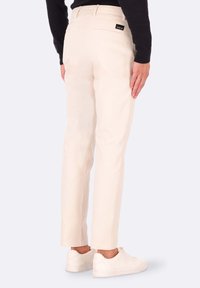 Pantaloni slim fit color crema con una texture liscia, tasche posteriori e una piccola etichetta nera del brand in vita. Indossati con sneakers bianche.