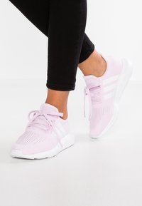 Osoba v světle růžových běžeckých botách s bílou podrážkou a pruhy, v černých těsných kalhotách, stojící na bílé ploše.