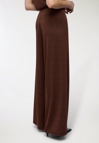 Jumpsuit marrone a fantasia con un design geometrico, gambe larghe e una texture liscia, completato da tacchi neri a punta.
