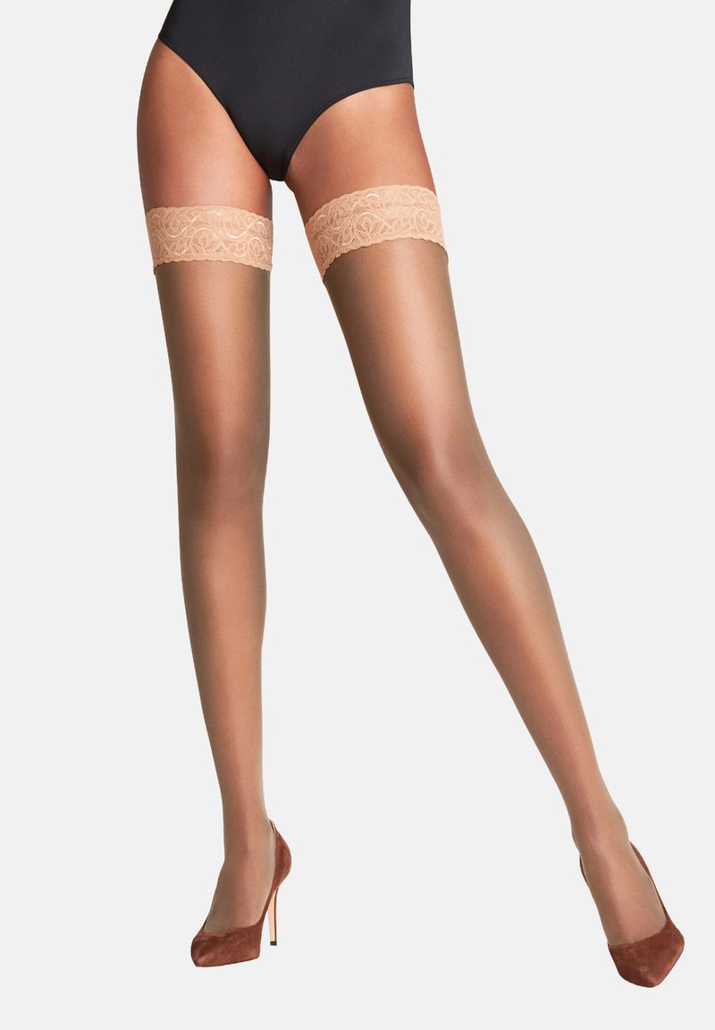 Collant color nude alti fino alla coscia con un top in pizzo decorativo. I collant hanno una texture liscia e lucida e sono abbinati a tacchi alti marroni.