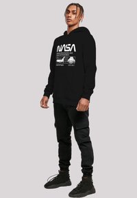 Black hooded sweatshirt featuring white NASA logo and text, with a front pocket and ribbed cuffs and hem. Worn with black cargo pants and boots.