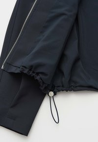 Manche de veste noire avec poignet élastique et cordon de serrage réglable, à côté d'une fermeture éclair argentée sur un fond de tissu lisse.