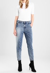 Nainen, jolla on päällään valkoinen tiukka paita, korkeavyötäröiset happopesu farkut ja mustat kärjet korkokengät, seisoo yksi käsi taskussa.