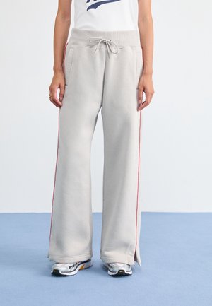 Personne portant un pantalon de survêtement ample gris clair avec des bandes rouges sur les côtés, une taille à cordon, et des chaussures de course blanches avec des accents noirs et jaunes.