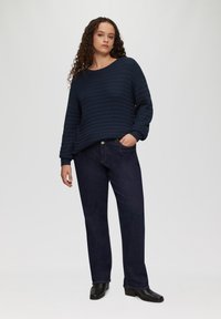 Marineblauer gerippter Pullover mit weiten Ärmeln, kombiniert mit dunkelblauen Jeans im geraden Schnitt. Das Model trägt schwarze Stiefeletten und hat die Hände in den Taschen.