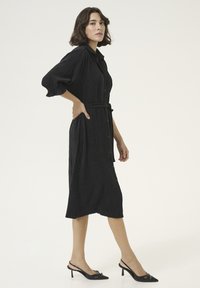Robe noire à boutons, avec une taille ceinturée, dotée d'un tissu texturé et de manches trois-quarts. Portée avec des talons noirs pointus.