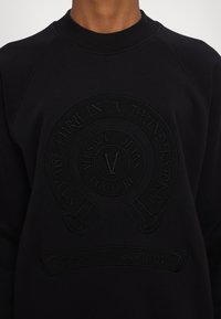 Sort sweatshirt med et hævet logodesign med tekst, rund halsudskæring og raglanærmer. Materialet ser tekstureret og blødt ud.