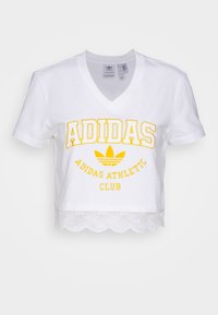 Vit beskuren t-shirt med v-ringning, med gul text "ADIDAS ATHLETIC CLUB" och spetsdetalj vid fållen. Mjuk bomullsmaterial.