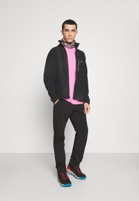Svart fleecejakke med glidelåslommer, rosa t-skjorte, svarte bukser og flerfargede sneakers med rosa detaljer. Nøytral bakgrunn.