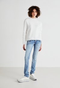 White ribbed sweater, light blue fitted jeans, and white sneakers with grey accents. Plain background highlights the outfit.