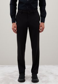 Uomo vestito con pantaloni neri, camicia a maniche lunghe nera e scarpe slip-on nere, in piedi su un pavimento tappezzato chiaro.