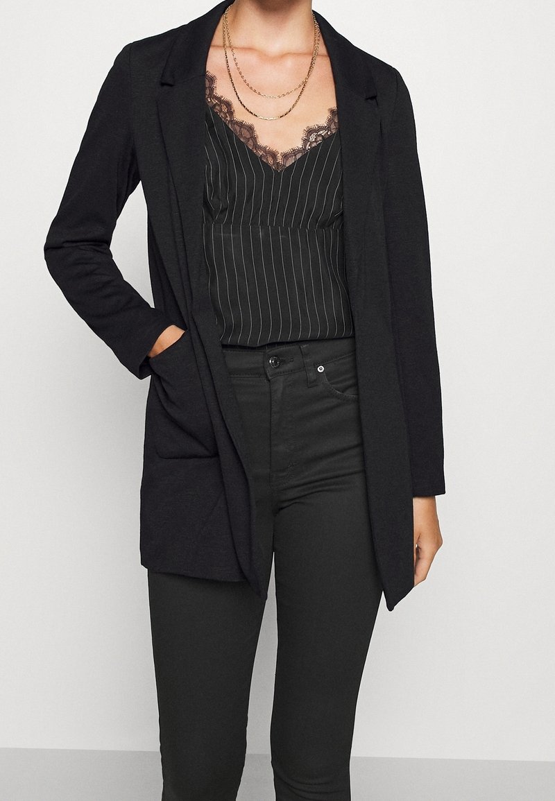 Czarny długi kardigan o luźnym kroju, z otwartym przodem i bocznymi kieszeniami, noszony na czarną bluzkę na ramiączkach w paski z koronkowym wykończeniem.