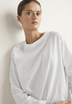 Junge Frau mit zurückgebundenem Haar, die ein schlichtes weißes Langarmshirt trägt und vor einer weißen Wand steht, nach vorn blickend.