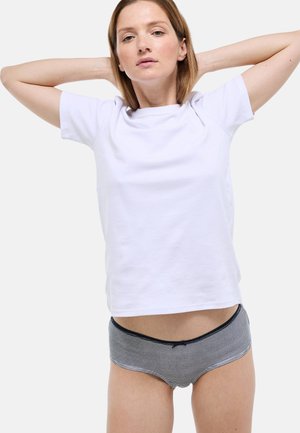 Femme aux cheveux châtain clair portant un t-shirt blanc uni et un sous-vêtement rayé noir et blanc, posant avec les mains derrière la tête sur un fond blanc.