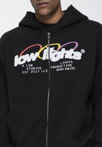 Sudadera negra con cremallera hecha de tela suave, con un logo gráfico colorido en el frente. Incluye capucha y bolsillos delanteros.