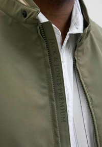 Olivově zelená bunda vyrobená z hladkého materiálu, s vysokým límcem, předním zipem a potištěným nápisem "JACK & JONES PREMIUM" na boku.