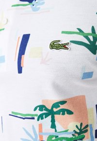 Polo blanc avec un motif abstrait coloré comprenant des palmiers verts, des formes géométriques et un logo crocodile brodé en vert.