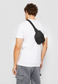 Sac à dos noir en tissu, doté d'une fermeture zippée et d'une bandoulière réglable, positionné dans le dos d'une personne portant un t-shirt blanc.