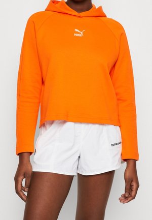 Personne portant un sweat à capuche Puma orange vif et un short de sport Puma blanc, se tenant avec les bras détendus devant un fond uni.