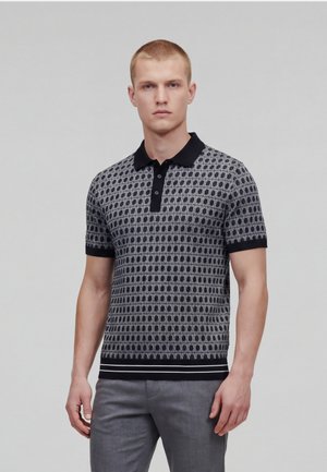 Junger Mann in einem kurzärmeligen Polohemd mit schwarzem und grauem Muster, schwarzem Kragen und grauer Hose, vor einem schlichten Hintergrund stehend.