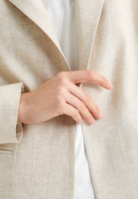 Ljust beige linblazer med en strukturerad slag. En hand justerar framknäppningen. Under finns en vit topp delvis synlig.