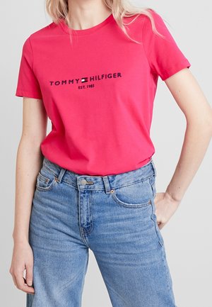 Kobieta w czerwonym t-shircie Tommy Hilfiger z logo i jasnoniebieskich, wysokich spodni jeansowych na jednolitym jasnym tle.