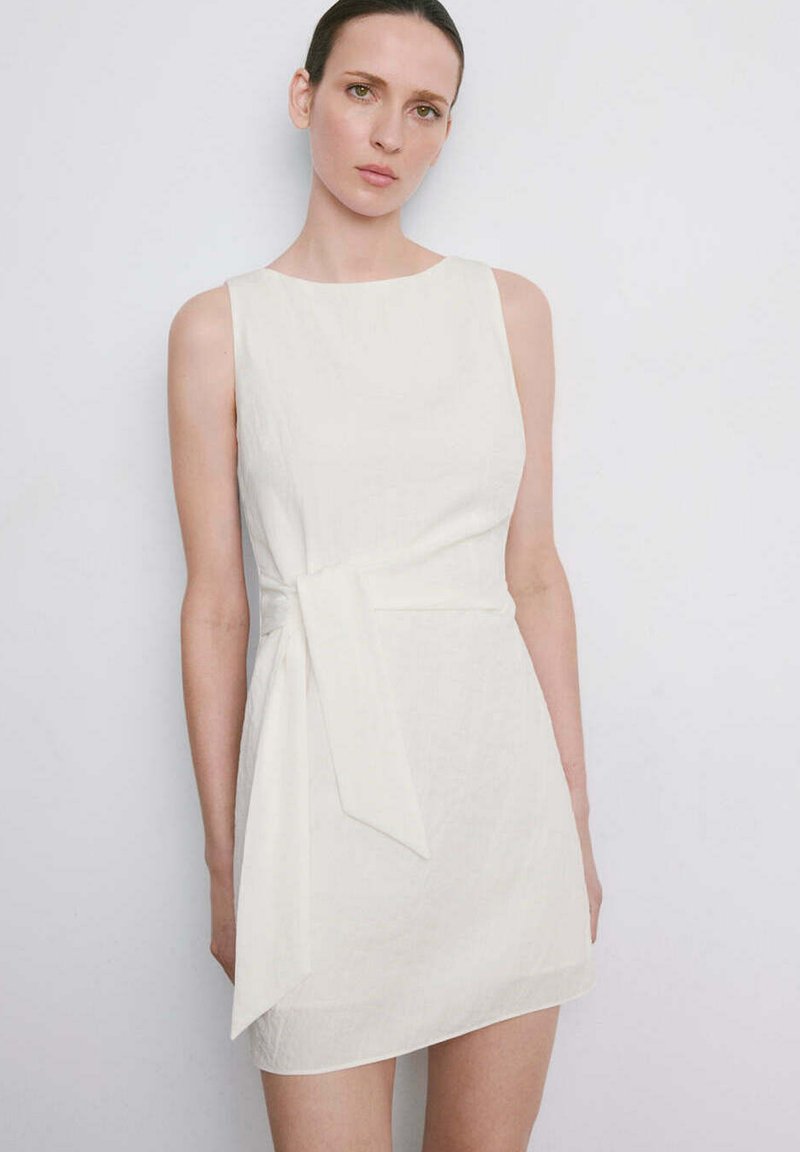Femme aux cheveux plaqués en arrière, portant une mini-robe blanche sans manches avec un détail de nœud sur le côté, debout devant un fond clair uni.