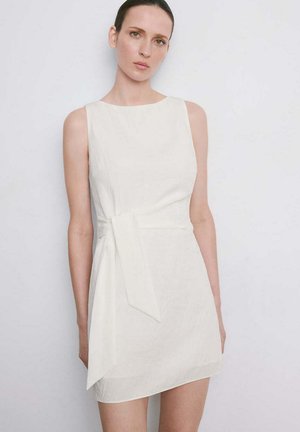 Mujer con el pelo peinado hacia atrás, vistiendo un mini vestido blanco sin mangas con un detalle de lazo lateral, de pie frente a un fondo claro y liso.