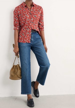 Personne portant une chemise boutonnée rouge à fleurs, un jean bleu à jambes larges, des mocassins noirs, et tenant un sac seau beige contre un mur blanc.