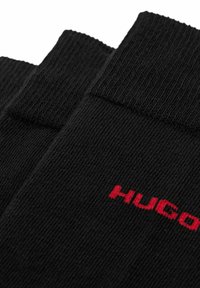 Chaussettes noires côtelées avec un logo "HUGO" rouge bien visible sur le côté. Texture douce avec un design classique de hauteur cheville.