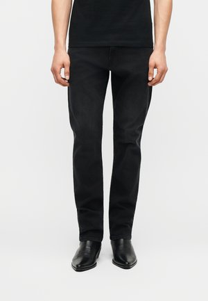 Άνδρας που φοράει στενά μαύρα τζιν και μαύρα δερμάτινα μποτάκια μέχρι τον αστράγαλο, στέκεται μπροστά σε απλό λευκό φόντο.