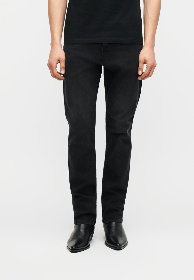 Άνδρας που φοράει στενά μαύρα τζιν και μαύρα δερμάτινα μποτάκια μέχρι τον αστράγαλο, στέκεται μπροστά σε απλό λευκό φόντο.