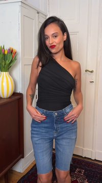 Γυναίκα με μακριά σκούρα μαλλιά που φοράει μαύρο τοπ ενός ώμου και τζιν σορτς μέχρι το γόνατο, στέκεται σε εσωτερικό χώρο κοντά σε βάζο με τουλίπες.